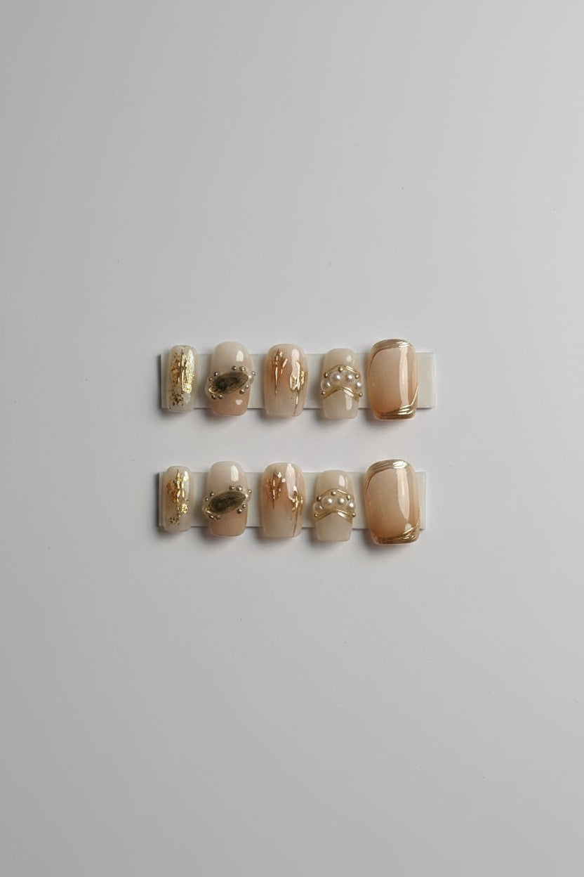 A hand-held card displaying 10 press-on nails from Dream Styles. The nails are beige and nude with various gold foil, glitter, and pearl embellishments.