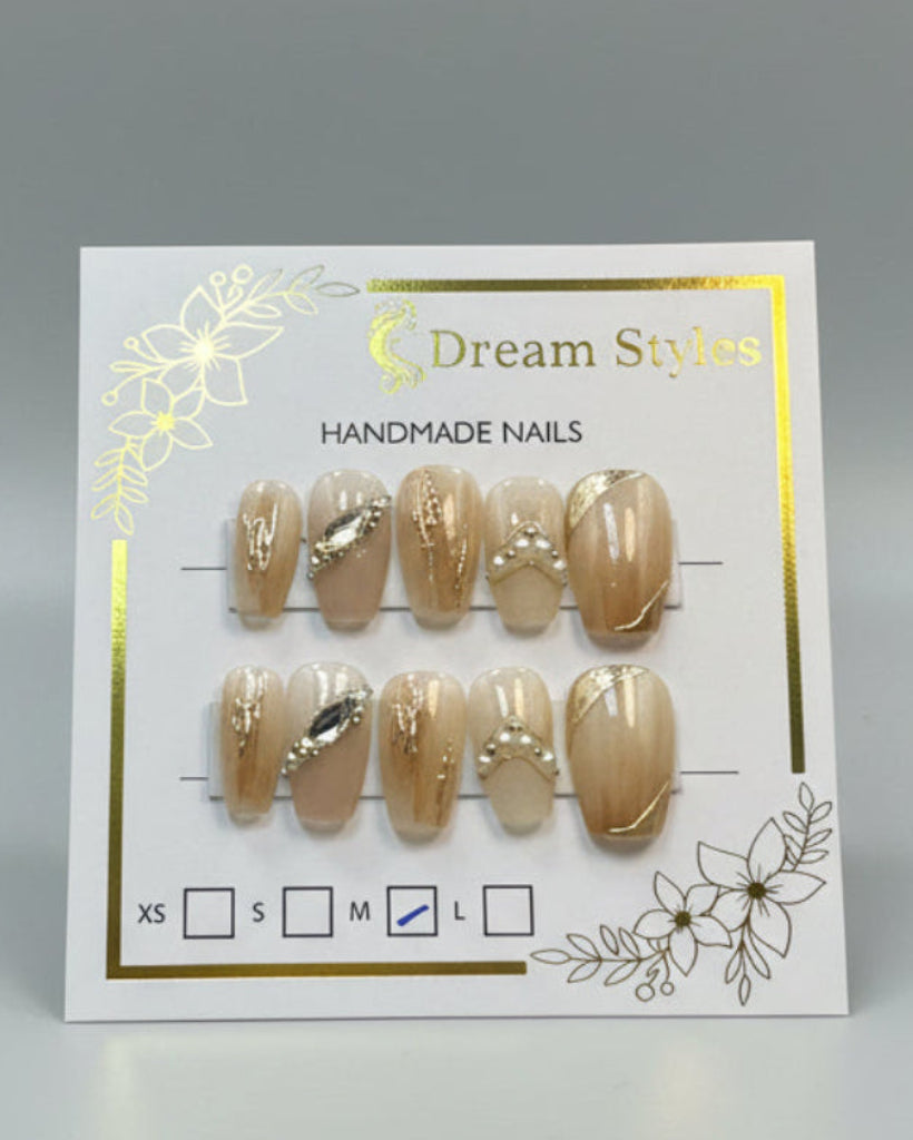 A set of 10 handmade press-on nails displayed on a branded card. The nails feature a glossy nude-beige base with various metallic gold and silver nail art designs, including rhinestones, curved foil, and pearl accents.